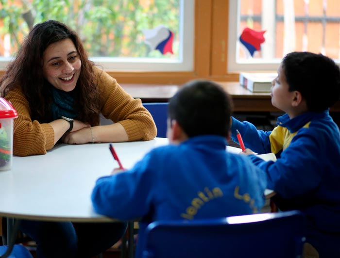 Tutora universitaria sonríe mientras asiste a niños en una sesión de tutoría en la Escuela Valentín Letelier, mejorando sus habilidades de lectura